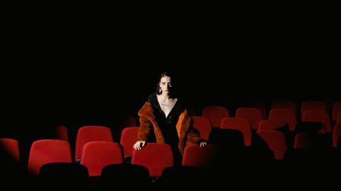 Woman in a fur-trimmed coat standing alone among rows of red seats in a dark cinema