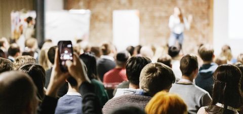 An audience seen from behind at a talk, with one person filming the speaker on a phone