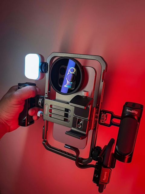 Hand holding a SmallRig phone cage with an LED light, lens filter and side handles against a red wall