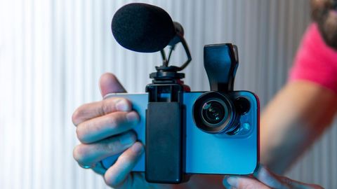 Hand holding a smartphone in a grip with a clip-on lens and a foam-covered microphone