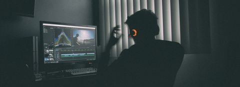 Video editor in headphones working on a timeline in desktop editing software, in a dark room with the blinds drawn