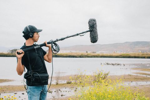 Sound recordist in headphones holding a boom microphone with a windshield beside a lake