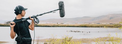 A sound recordist wearing a black cap, headphones, and black shirt stands outdoors holding a long boom pole with a large fluffy windscreen (dead cat) microphone, pointing it toward a lake with birds on the water and hills behind.